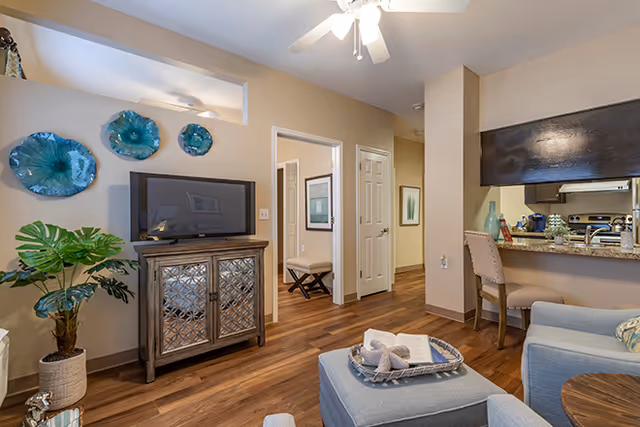 A cozy living room area with a flat-screen TV on a decorative wooden cabinet, blue glass wall art above the TV, a potted plant to the left, and a light blue armchair with a matching ottoman in the foreground. The room has wood flooring and beige walls. In the background, there is a small dining area with a chair and a kitchen with granite countertops and dark cabinets.