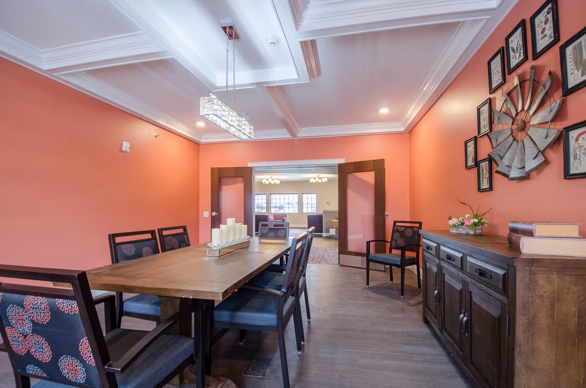 A dining room with coral-colored walls, a wooden dining table surrounded by six chairs with dark frames and patterned cushions. On the table is a rectangular tray holding several white pillar candles. To the right is a wooden sideboard with books and a small flower arrangement. Above the sideboard is a decorative metal windmill surrounded by framed botanical prints. The room has a coffered ceiling with a modern rectangular chandelier. Double wooden doors with frosted glass panels open to a bright living area with large windows and seating.