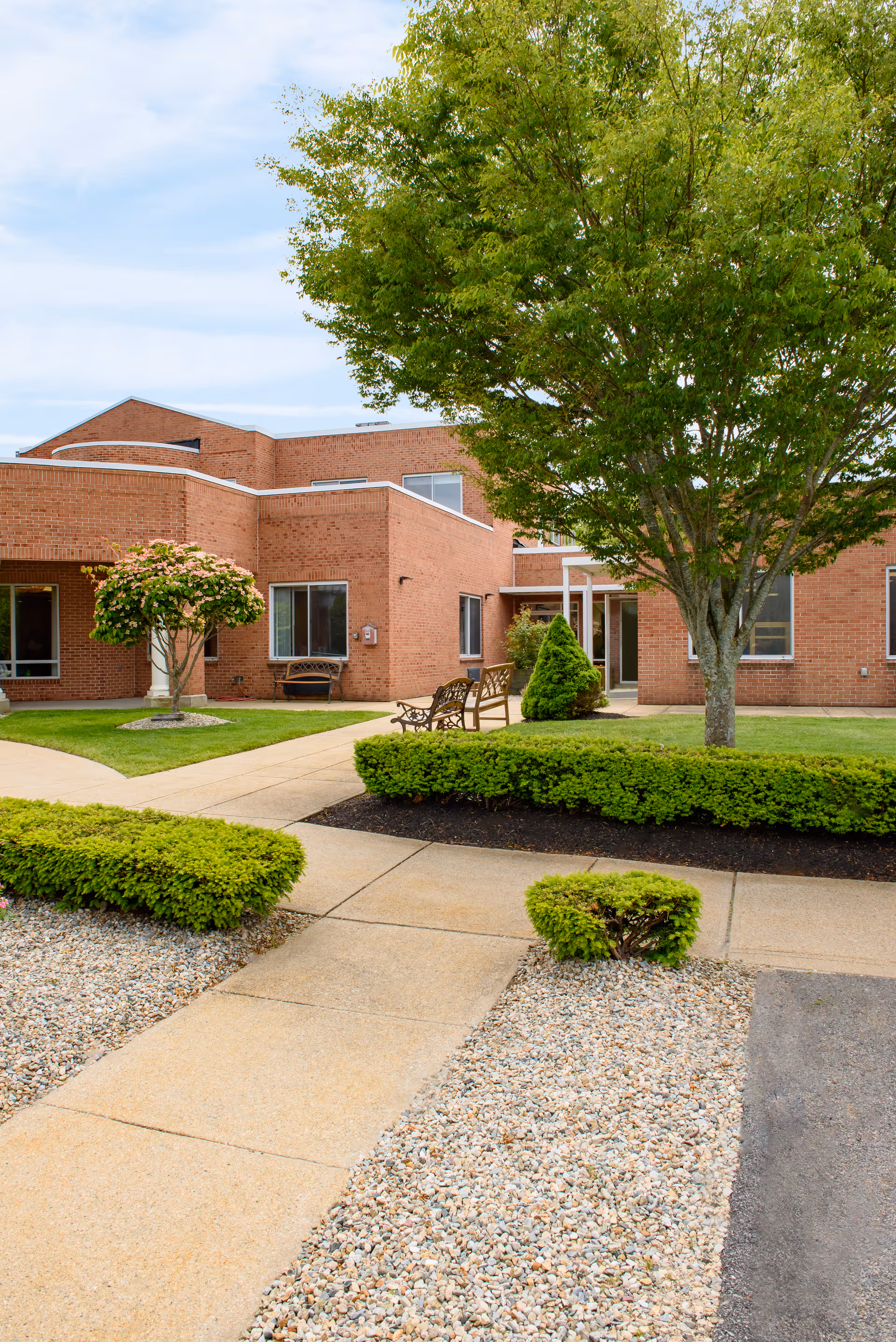 Outdoor view of a brick building with a well-maintained garden area featuring green bushes, a large tree, a smaller flowering tree, and benches along a paved walkway under a partly cloudy sky.