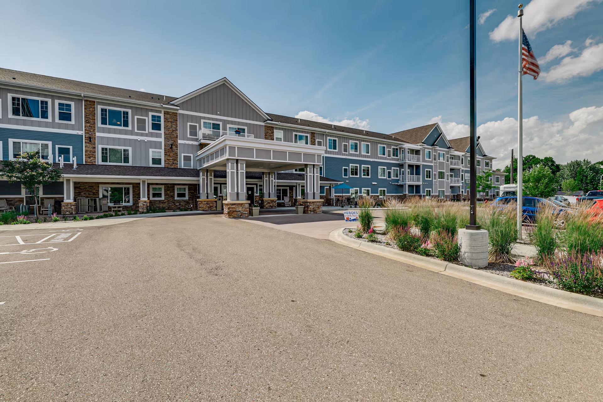 Exterior view of a multi-story senior living facility building with a covered entrance driveway. The building features a combination of gray and blue siding with white trim and stone accents. There is a landscaped area with tall grasses and flowers near the entrance, an American flag on a flagpole, and a parking lot with several cars. The sky is clear with a few clouds.