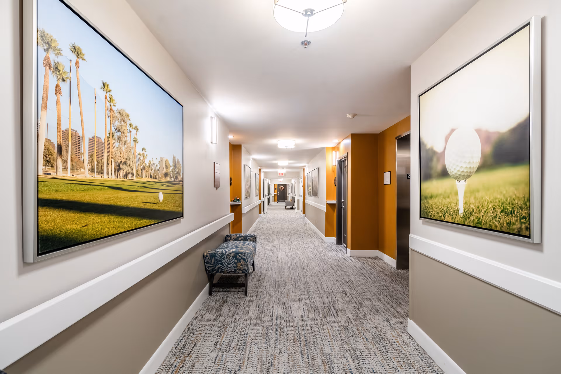 A well-lit indoor hallway with carpeted flooring and beige and white walls. The hallway features framed pictures of a golf course and a close-up of a golf ball on the grass. There is a patterned bench along the left wall and an elevator on the right side. The hallway extends into the distance with additional seating and artwork visible.