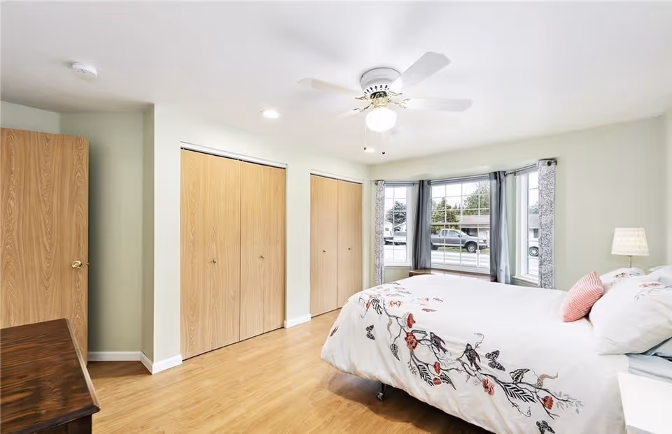 Bright bedroom with a large bed covered in white bedding with floral patterns, a ceiling fan with light, wooden floor, two large closets with wooden doors, a window with curtains showing a view of parked cars outside, and a small bedside lamp.