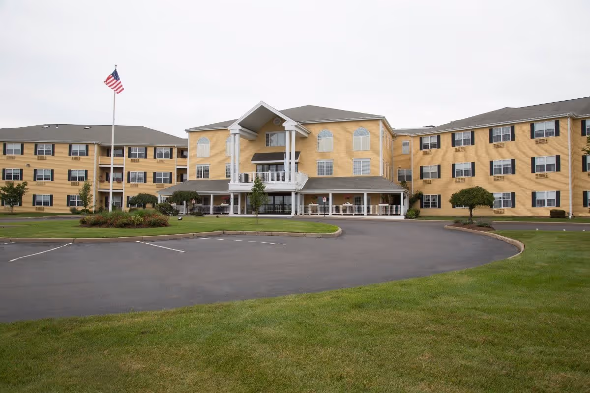 Exterior view of a large, three-story senior living facility building with yellow siding and white trim. The building has multiple windows with black shutters and a covered entrance supported by white columns. There is a circular driveway and a flagpole with an American flag in front, surrounded by well-maintained green lawns and small trees.
