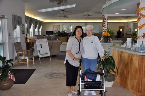 Two women standing inside a senior living facility. One woman is elderly and using a walker, while the other woman stands beside her with an arm around her shoulder. The interior features a reception desk, chairs, plants, and a signboard in the background.