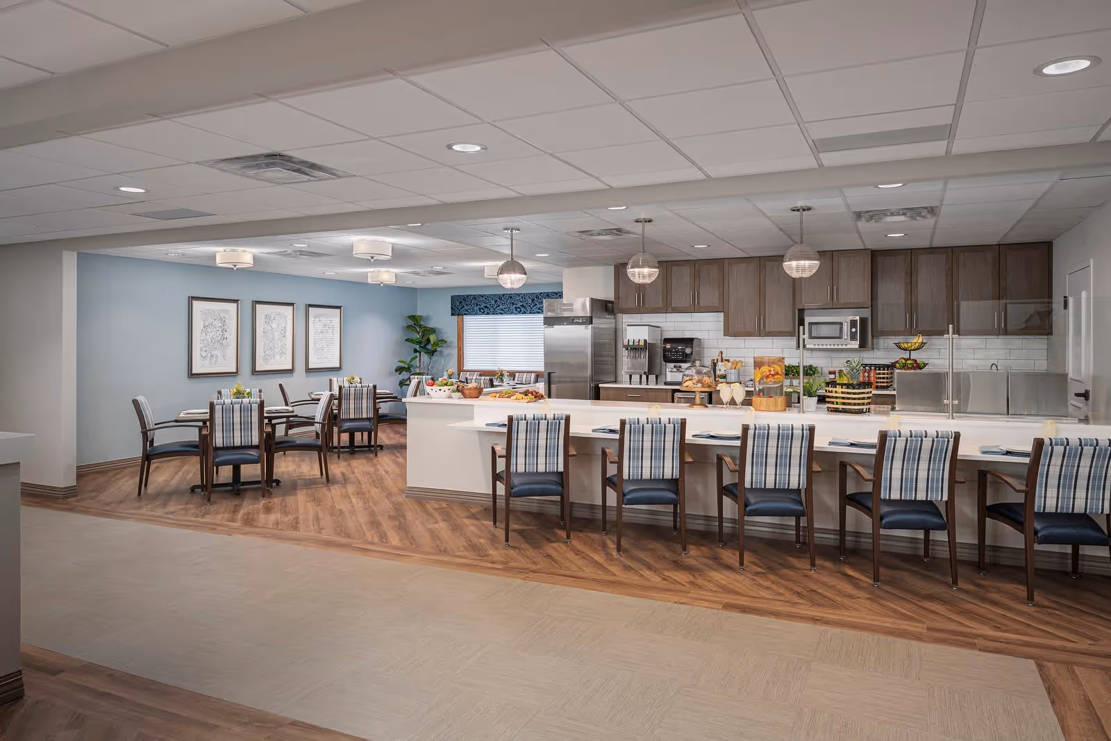 Communal dining room and open kitchen with a long island lined with plaid-upholstered chairs and round dining tables in the background.