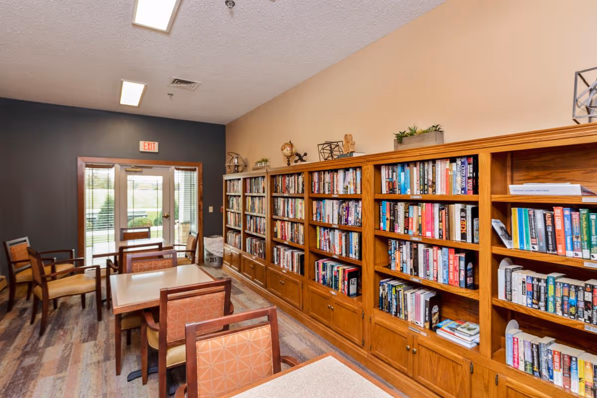 Interior of a senior living facility room with wooden bookshelves filled with books along one wall, several tables and chairs arranged for reading or socializing, and a glass door leading outside with an exit sign above it.
