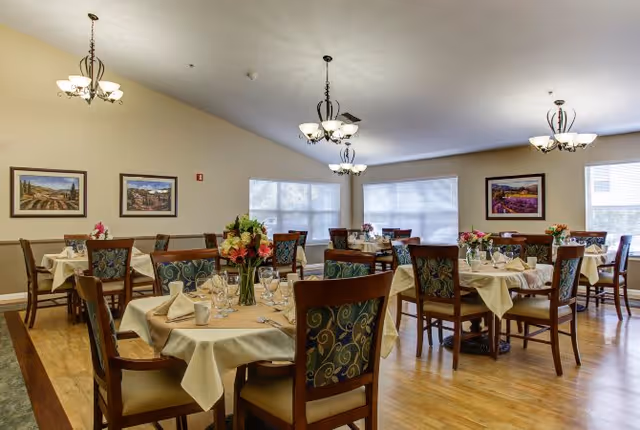 A bright dining room with multiple round tables covered with beige tablecloths, each set with glassware, napkins, and floral centerpieces. The room has wooden chairs with patterned upholstery, light wood flooring, beige walls, framed landscape paintings, and large windows with white blinds. Elegant chandeliers hang from the ceiling providing warm lighting.