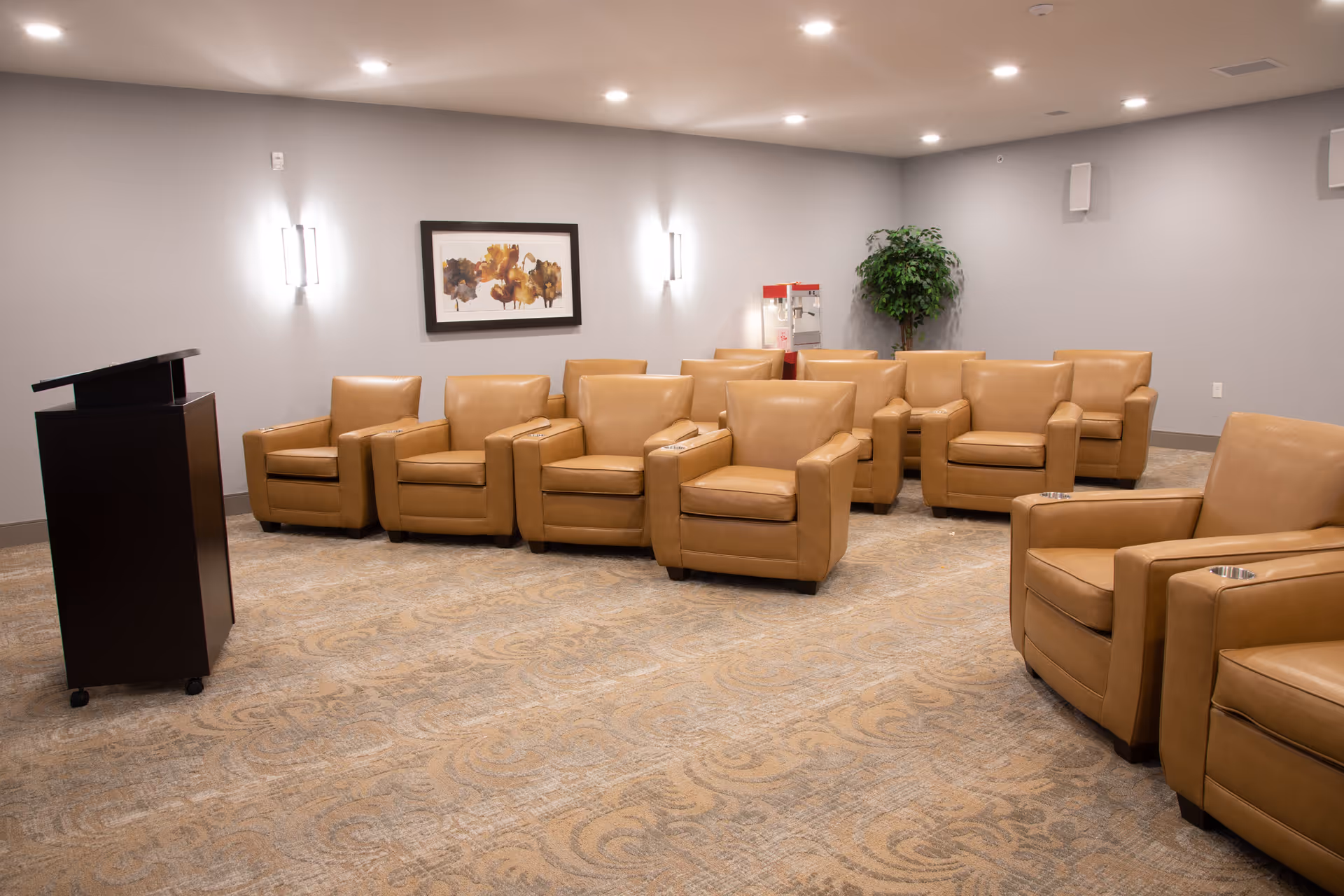 A small conference or presentation room with two rows of tan leather armchairs facing a dark wooden podium. The room has light gray walls, a patterned carpet, wall-mounted lights, a framed abstract painting, a popcorn machine, and a potted plant in the corner.