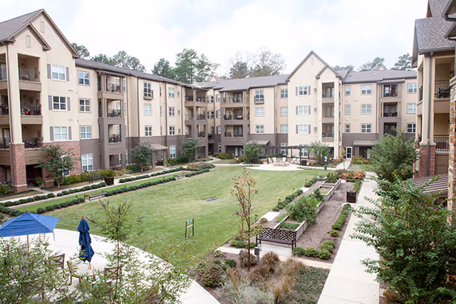 View of a large courtyard in the center of a multi-story senior living facility with well-maintained green lawn, benches, garden beds, and outdoor seating areas surrounded by beige and brown buildings with balconies.