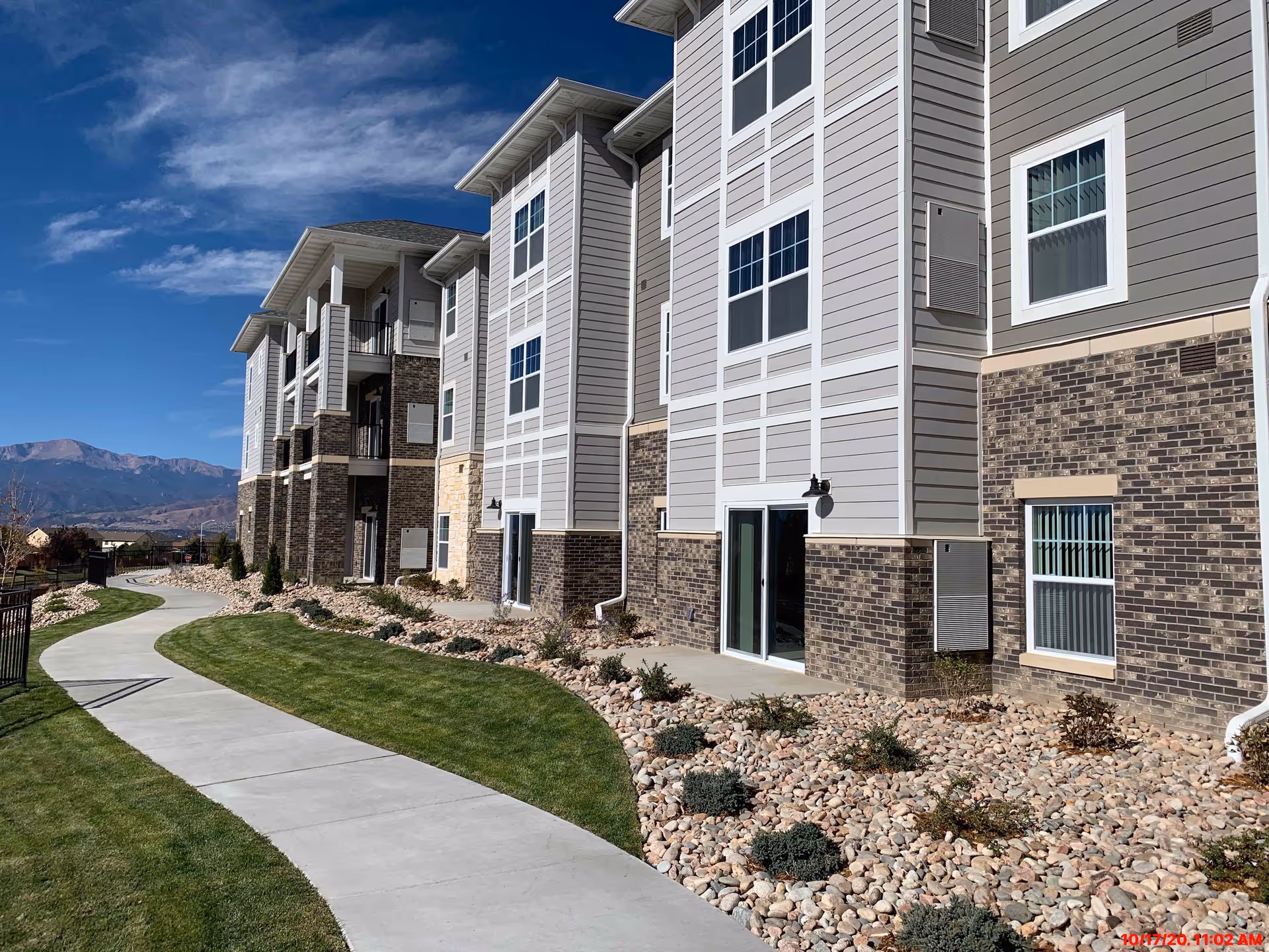 Exterior of a three-story retirement building with a curved sidewalk, rock landscaping and mountains in the background.