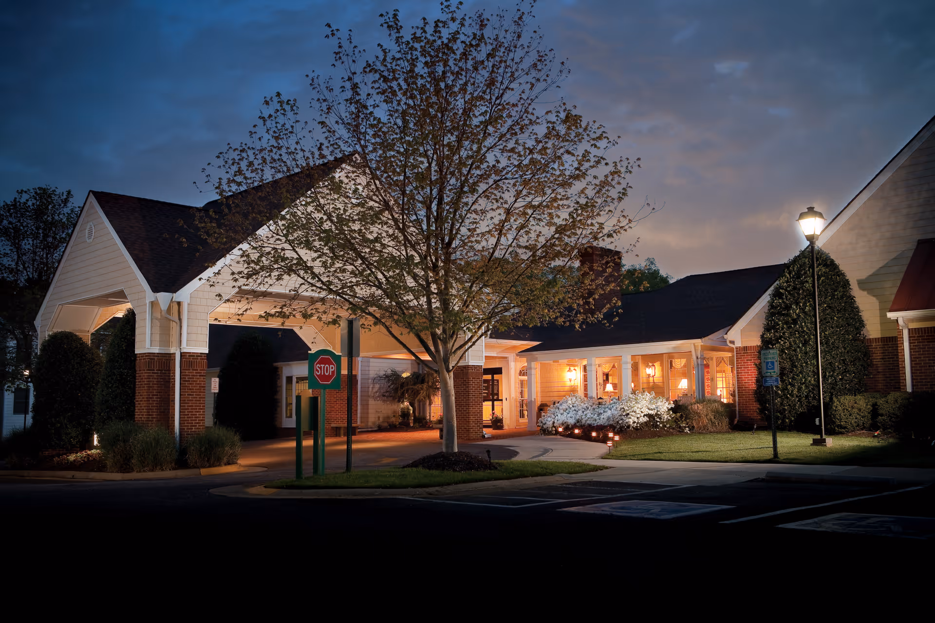 Lit entrance canopy and front façade of a retirement community at dusk, with a stop sign, tree, and landscaping.
