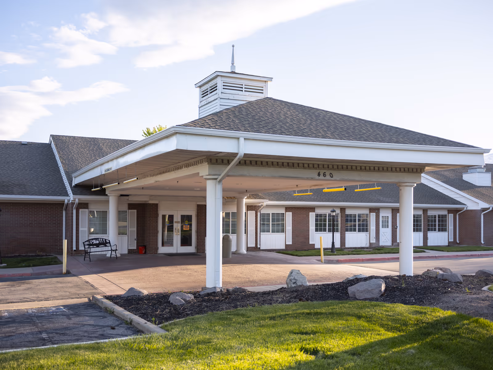 Exterior view of Monument Healthcare Bountiful Skilled Nursing facility showing the main entrance with a covered drop-off area supported by white columns, brick walls, multiple windows, and a bench near the entrance.