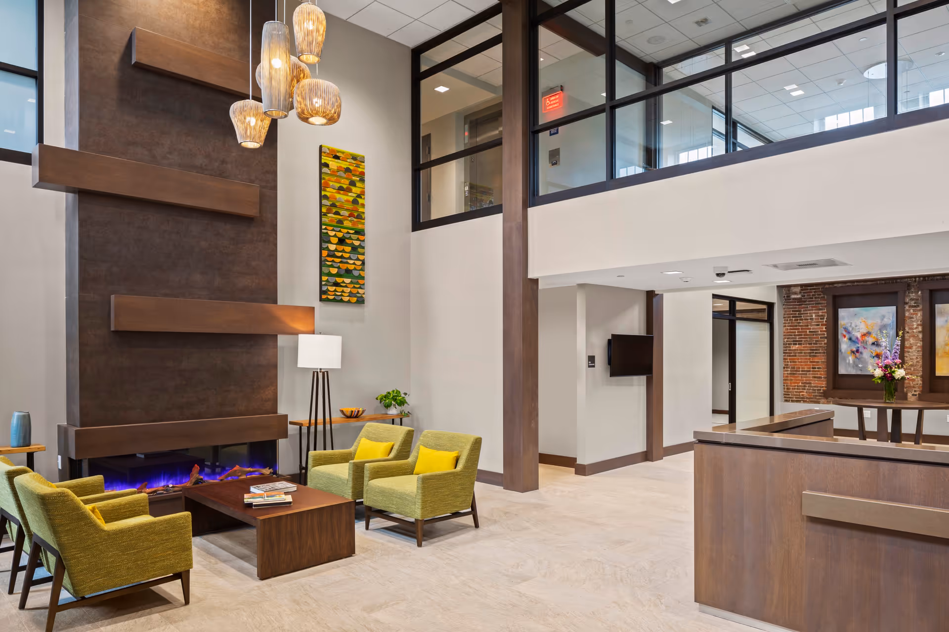 A modern and spacious living room area with a tall dark brown fireplace featuring horizontal wooden accents and a blue flame. Four green armchairs with yellow cushions surround a wooden coffee table with books on it. A floor lamp and a small side table with a plant are next to the fireplace. Above the fireplace hangs a colorful abstract wall art. The room has large windows on the upper level, beige walls, and light-colored flooring. In the background, there is a hallway with a mounted TV and a table with flower arrangements and framed artwork on the brick wall.