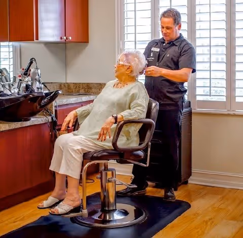 An elderly woman is seated in a salon chair while a hairstylist stands behind her, styling her hair. The setting appears to be a well-lit room with large windows covered by white shutters and wooden flooring. There are salon sinks and cabinets visible on the left side.