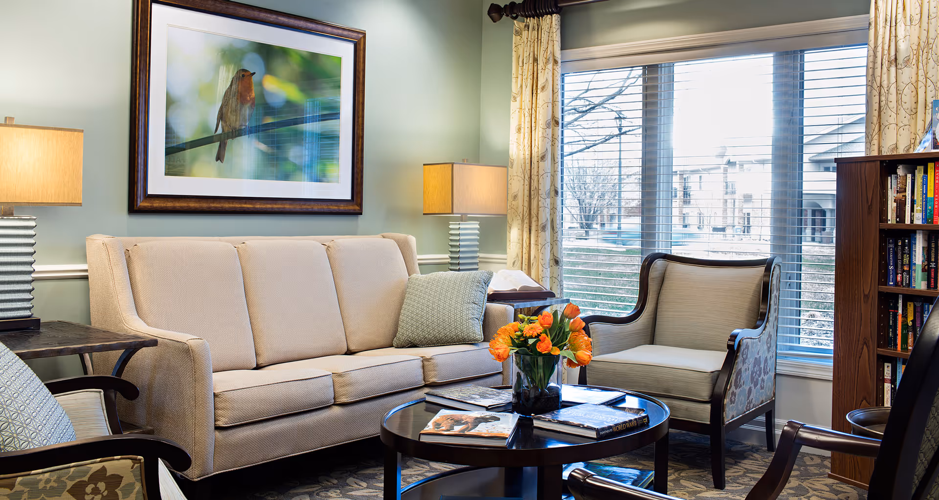 Bright living room with a beige sofa and armchairs around a round coffee table topped with a vase of orange tulips, lamps, artwork, and a large window with blinds.