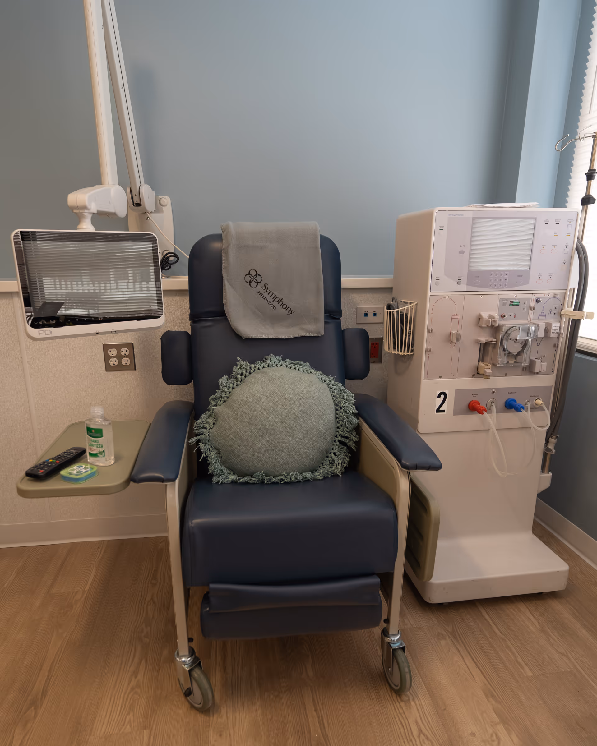 A medical treatment room featuring a blue reclining chair with a round green cushion and a gray blanket draped over the backrest. To the right of the chair is a dialysis machine with tubes and controls. To the left is a small attached table holding a remote control, a bottle of hand sanitizer, and a small container. The room has light blue walls and wood flooring.