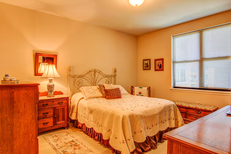 A cozy bedroom with a double bed covered in a cream-colored quilt with floral patterns. The room features wooden furniture including a dresser, nightstand with a decorative lamp, and a chest under the window. There are framed pictures on the walls and a window with blinds letting in natural light.