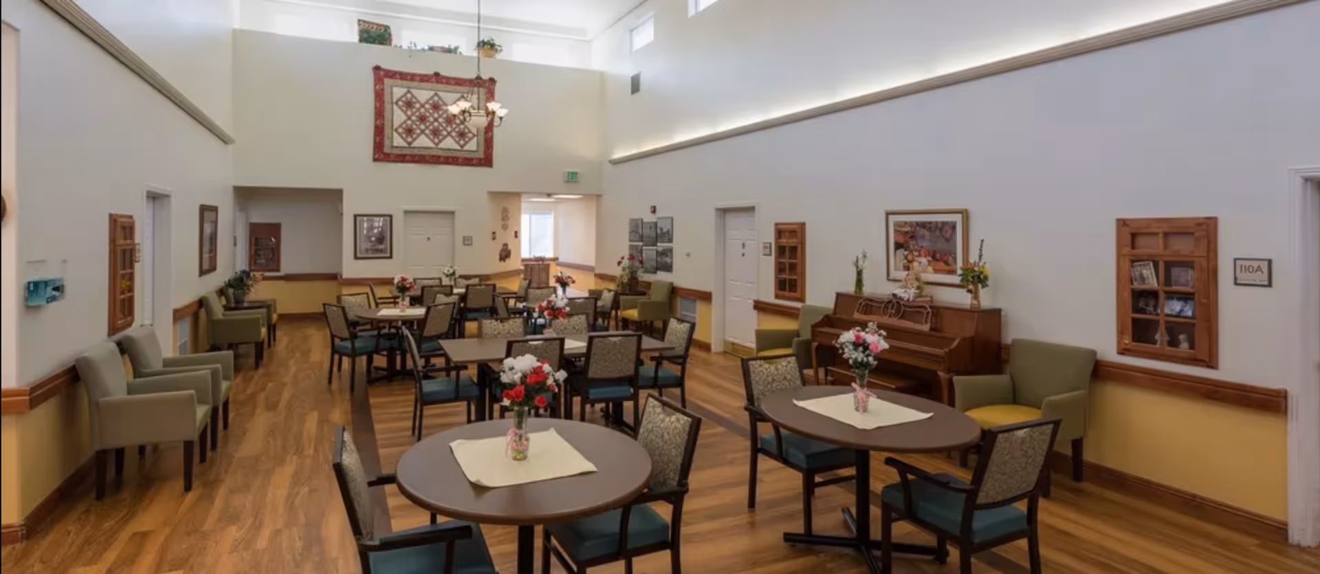 Spacious communal dining/activity room with round tables and chairs, floral centerpieces, a piano, and high ceilings.