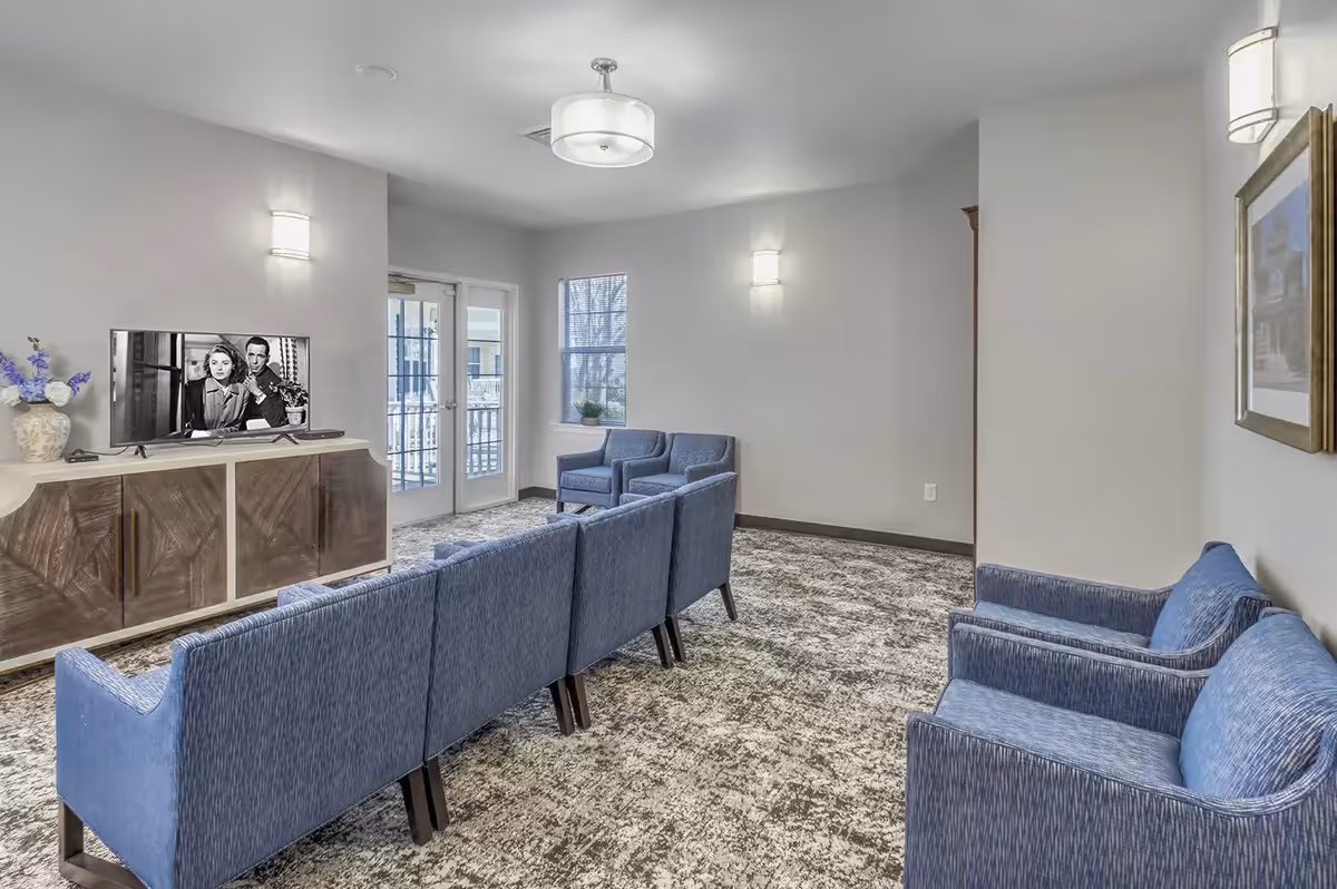 A modern living room with blue upholstered chairs and sofas arranged facing a flat-screen TV on a wooden cabinet. The room has light gray walls, carpeted floor, wall-mounted lights, a ceiling light fixture, and a window and glass door leading outside.