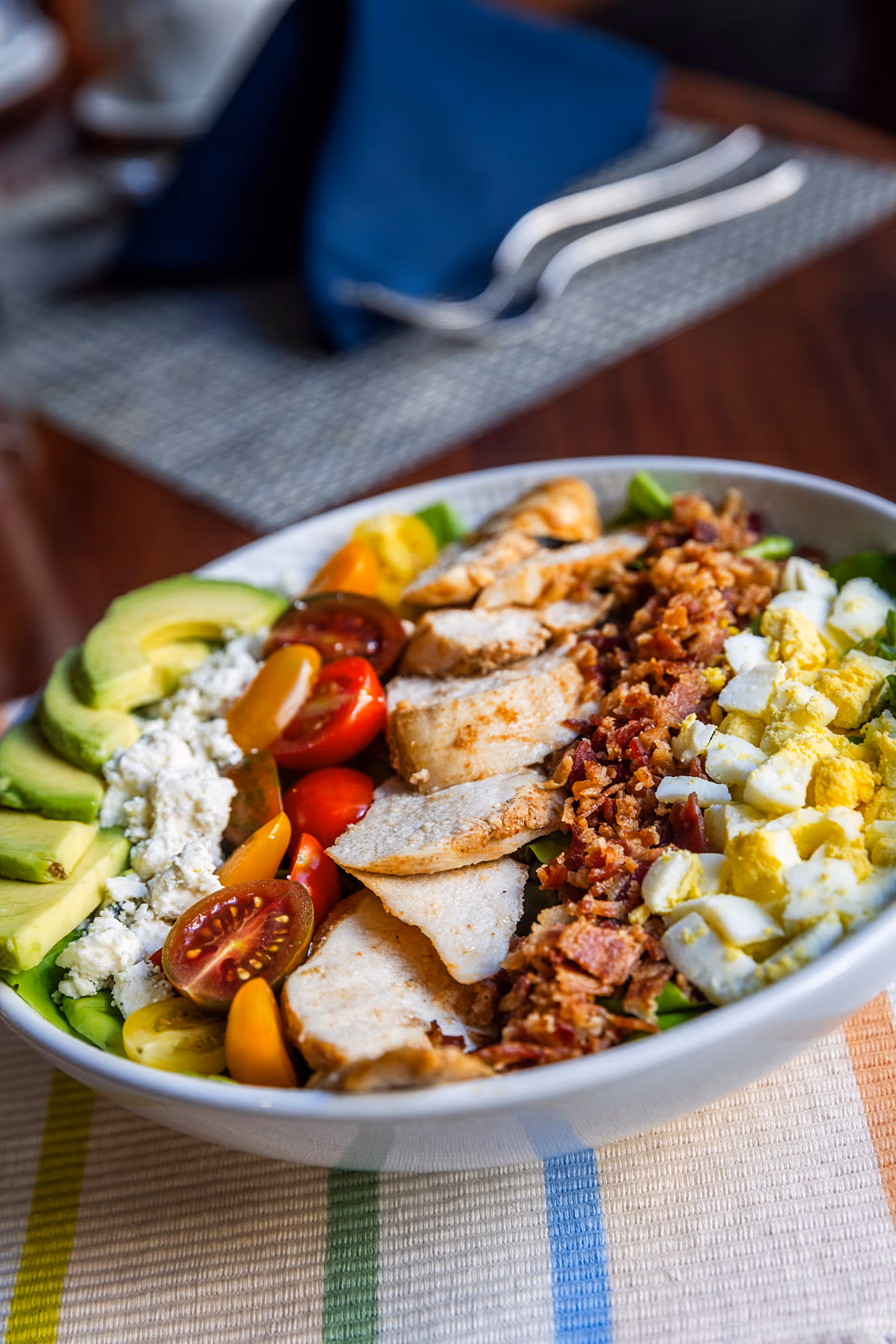 Close-up of a large Cobb-style salad bowl with sliced grilled chicken, avocado, cherry tomatoes, bacon, chopped hard-boiled egg and crumbled cheese on a table.