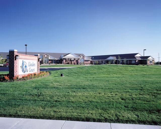Wide view of the exterior of Linden Court facility with a large green lawn in the foreground and a clear blue sky. A brick sign with the name 'Linden Court' is visible on the left side near the sidewalk.