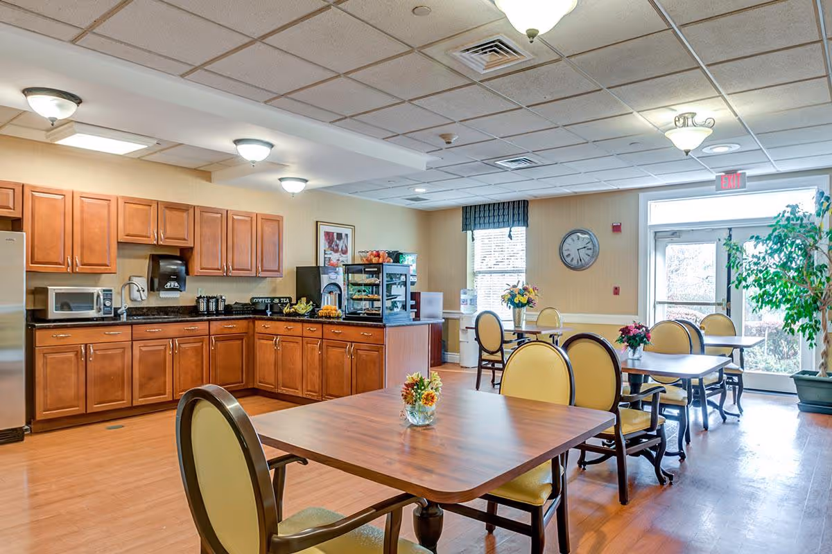 A bright dining area with wooden tables and yellow cushioned chairs. The room features a kitchenette with wooden cabinets, a microwave, coffee and tea station, and a display case with snacks. There are windows with blinds, a clock on the wall, and a glass door leading outside. Potted plants and small flower arrangements decorate the space.