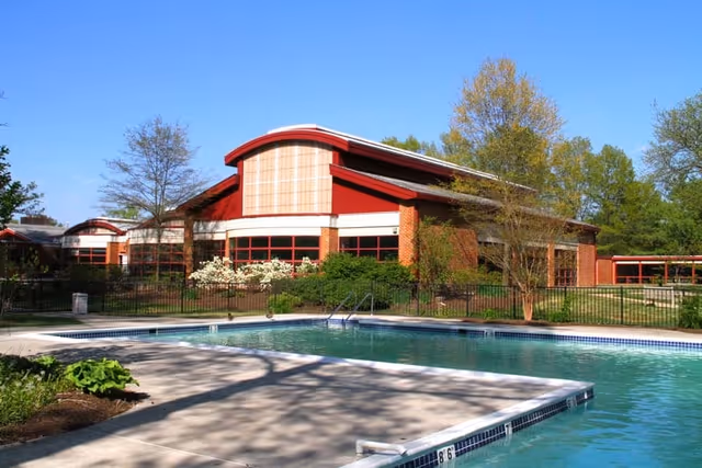 Outdoor swimming pool with clear water in front of a red brick building with large windows and a curved roof, surrounded by trees and greenery under a clear blue sky.