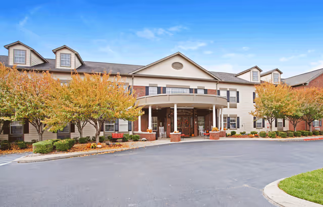Front exterior view of The Carriages of Rivergate senior living facility with a covered entrance, brick and beige siding, several windows, and trees with autumn foliage lining the driveway.