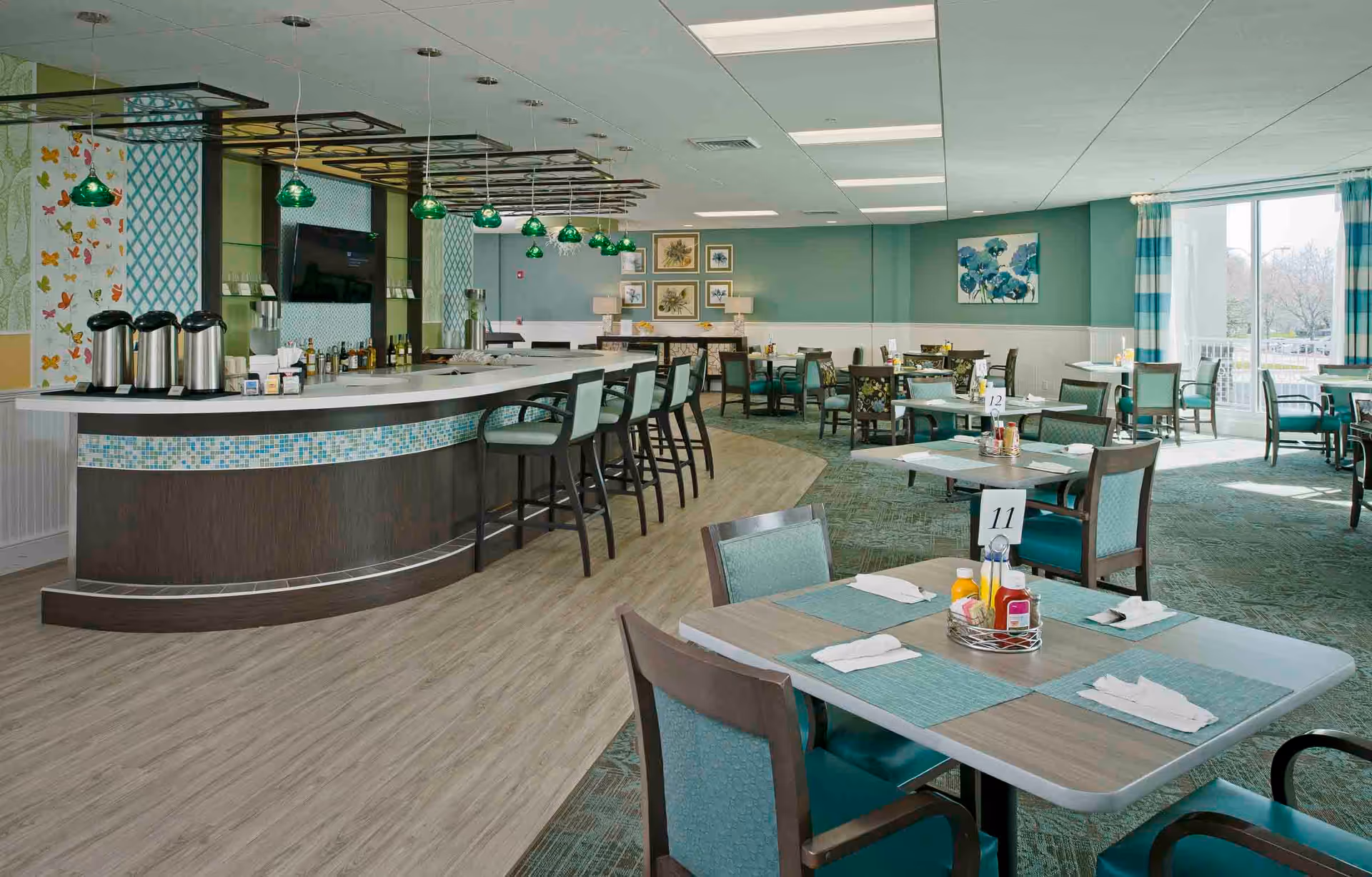 A bright and spacious dining area in a senior living facility with multiple tables and chairs arranged neatly. The tables have placemats, napkins, and condiment holders. There is a curved bar counter with several high chairs and green pendant lights hanging above it. The walls are decorated with framed artwork and patterned wallpaper, and large windows with striped curtains allow natural light to fill the room.