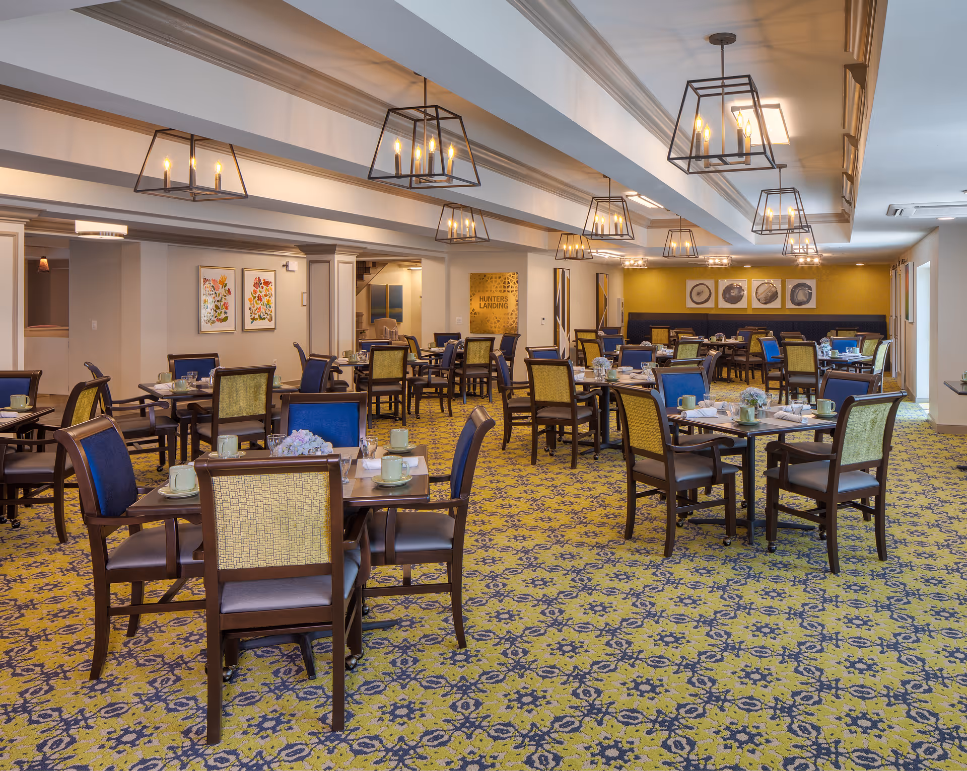 A spacious dining room with multiple tables set with cups, plates, and napkins. The room features patterned yellow and blue carpet, wooden chairs with blue and yellow upholstery, modern hanging light fixtures, and framed artwork on the walls. The space is well-lit with natural light coming through windows on the right side.