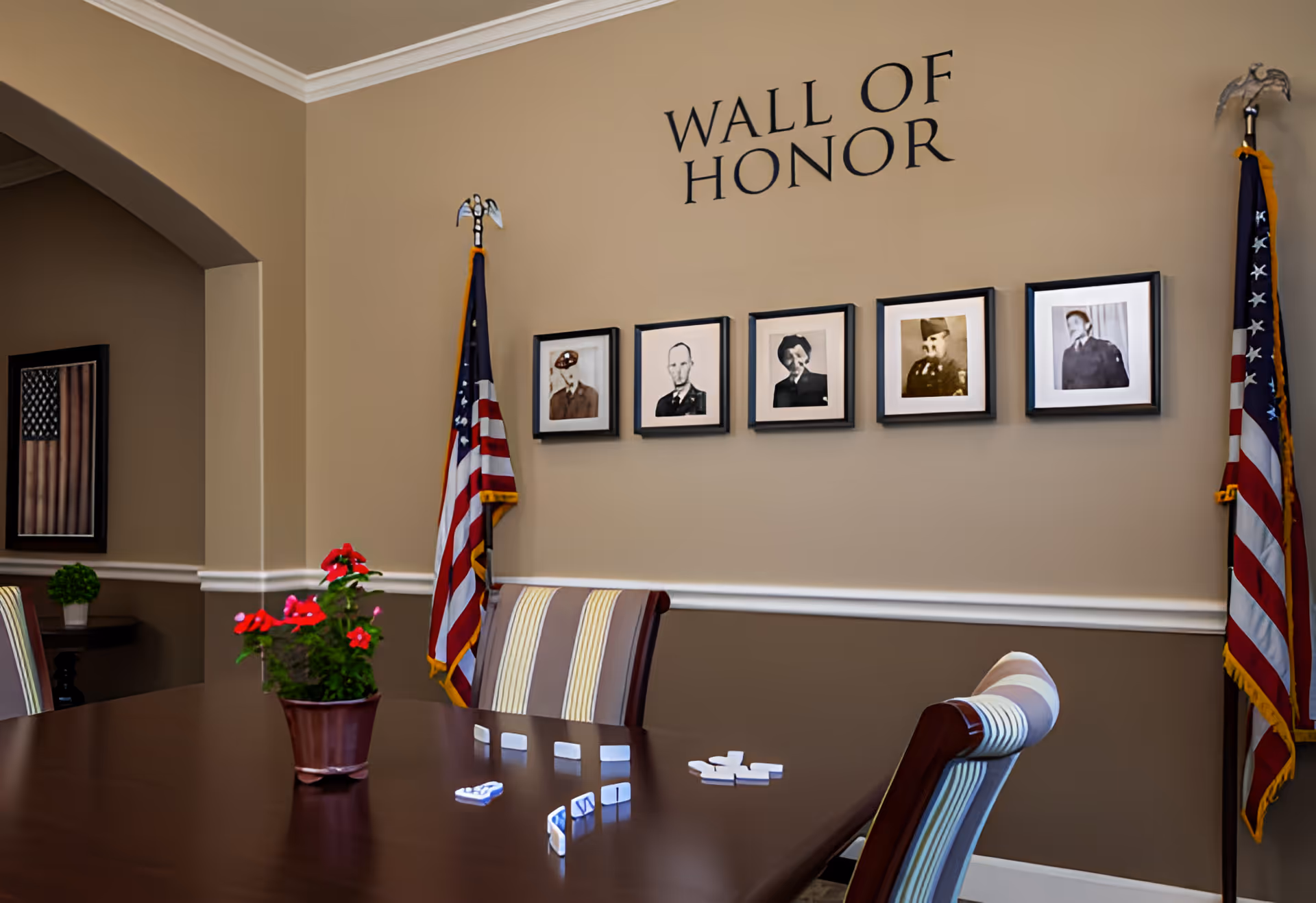 A room with a wooden table and striped chairs, featuring a potted plant with red flowers on the table. On the wall above the table is a 'Wall of Honor' display with five framed black and white portraits of military personnel. Two American flags stand on either side of the portraits. Another framed American flag is visible on a side wall.