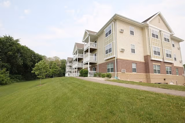 Exterior view of a multi-story senior apartment building with beige siding and brick accents, surrounded by a grassy lawn and trees under a partly cloudy sky.