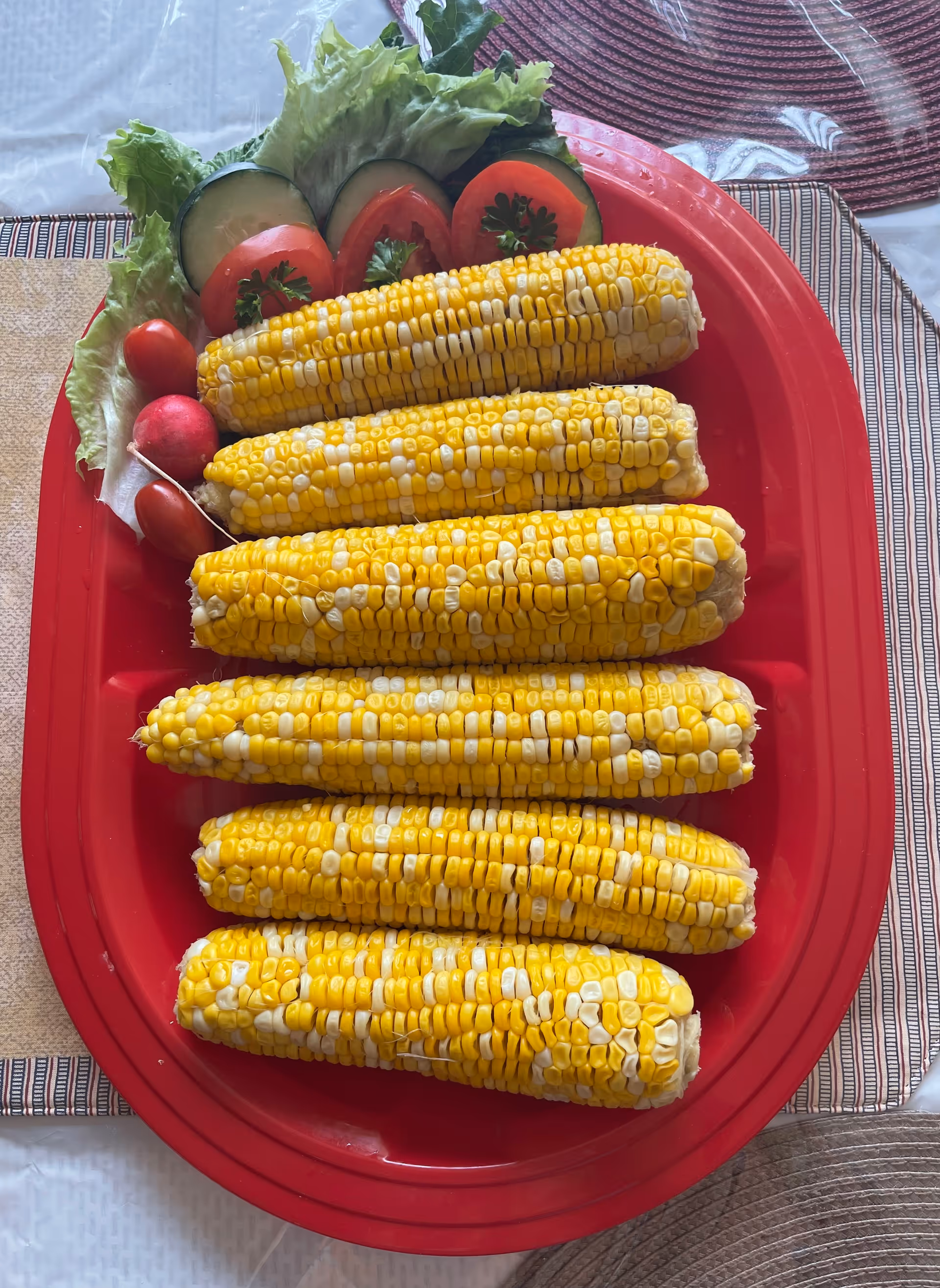 A red platter with six ears of yellow and white corn on the cob, garnished with lettuce, sliced cucumbers, tomato slices, radishes, and grape tomatoes, placed on a table with placemats.