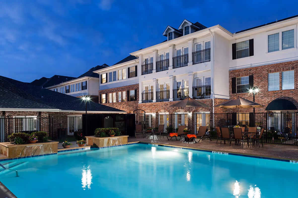 Outdoor swimming pool area at dusk with lounge chairs, tables with umbrellas, and a multi-story brick and white building in the background.