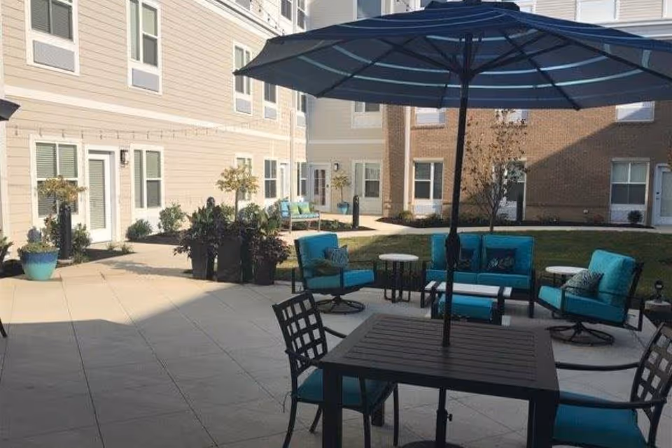 Outdoor patio area at Traditions at North Willow with blue cushioned chairs, a table with an umbrella, potted plants, and surrounding residential building walls with windows and doors.