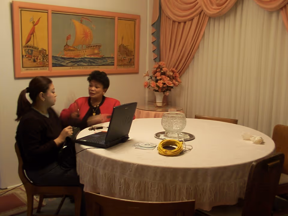 Two women sitting at a round dining table covered with a white tablecloth. One woman is using a laptop while the other is engaged in conversation. The room has peach-colored curtains, a floral arrangement on a side table, and a framed painting of a ship on the wall.