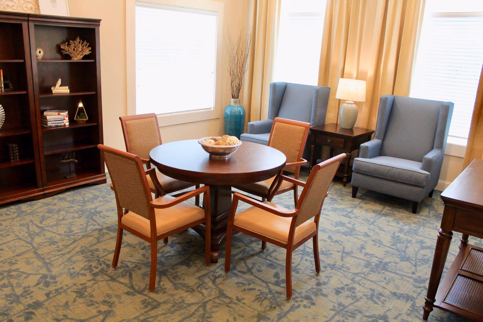 A cozy sitting area in Scotland House featuring a round wooden table surrounded by four cushioned wooden chairs. Behind the table are two blue upholstered armchairs with a small wooden side table and lamp between them. A tall blue vase with decorative branches is placed near the window. A wooden bookshelf with decorative items and books is visible on the left side. The room has large windows with beige curtains and a patterned carpet.