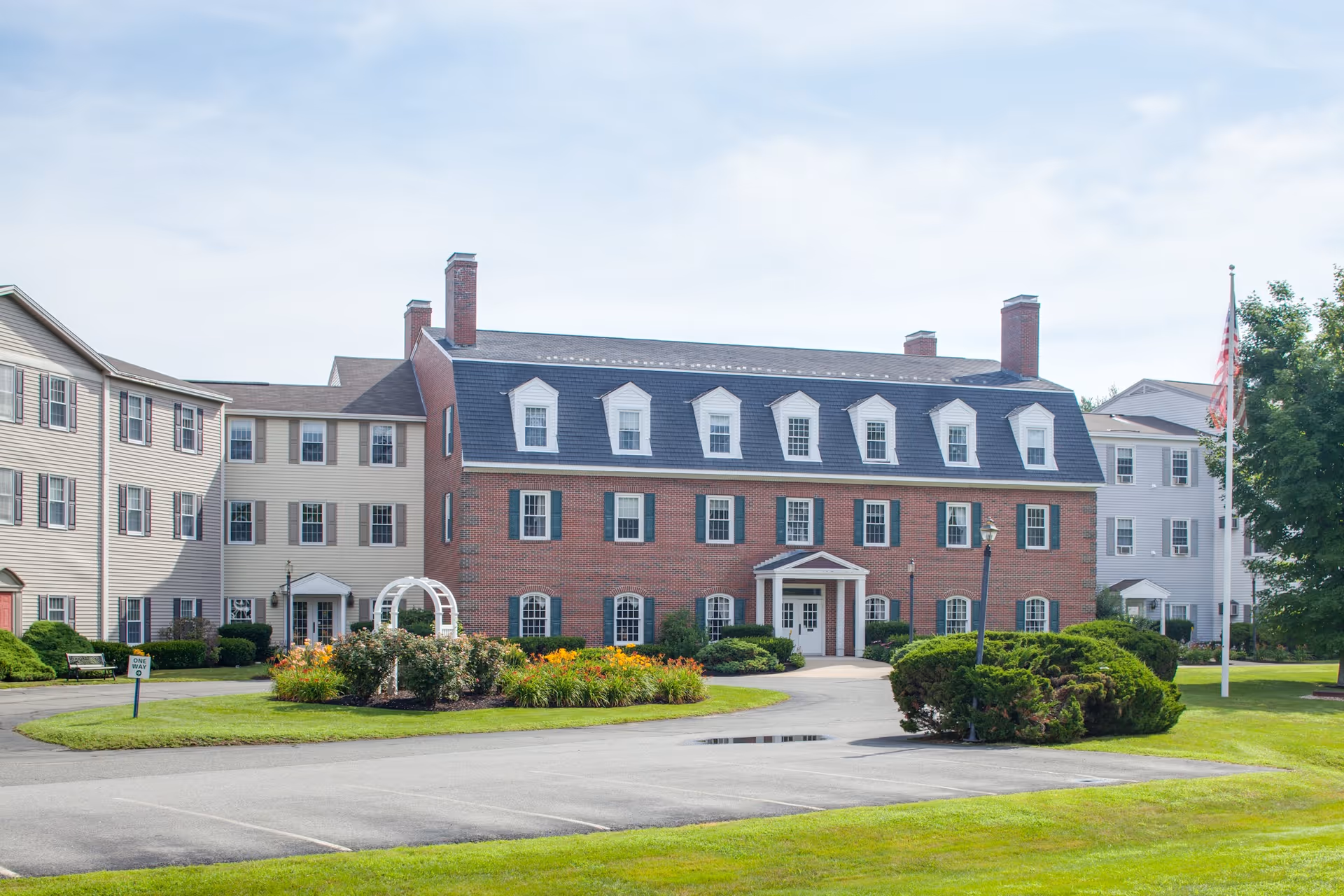 Front exterior of a three-story senior living building with a central brick section, dormer windows, landscaped circular driveway and an American flag.