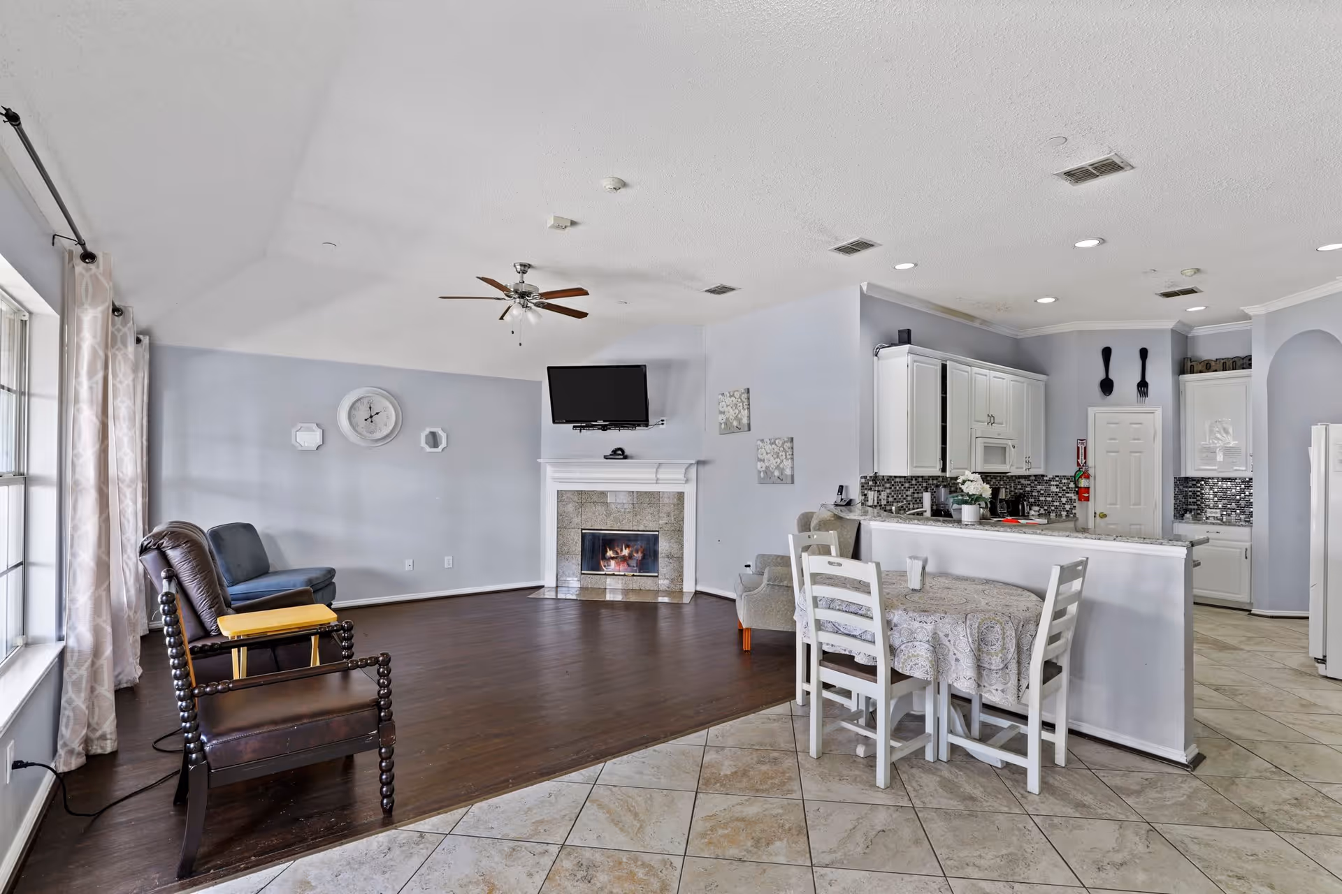 A spacious senior living common area featuring a seating area with two chairs near large windows with curtains, a fireplace with a mounted TV above it, and a kitchen with white cabinets, a tiled backsplash, and a small dining table with four chairs covered with a patterned tablecloth.