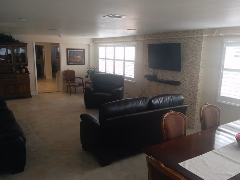 A living room area with black leather sofas and armchairs facing a wall-mounted flat screen TV. The TV is mounted on a textured stone wall with a small shelf below it holding electronic devices. There are two windows with white blinds allowing natural light into the room. To the right, part of a wooden dining table with chairs is visible. On the left side, there is a wooden cabinet and a framed painting on the wall near an open doorway.