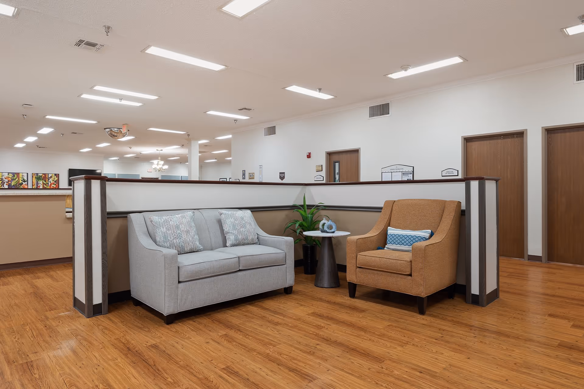 A seating area in a nursing and rehabilitation center featuring a light gray loveseat with two patterned pillows, a brown armchair with a blue and white pillow, and a small round table with a plant and decorative item. The area has wood flooring, white walls, and several wooden doors in the background.