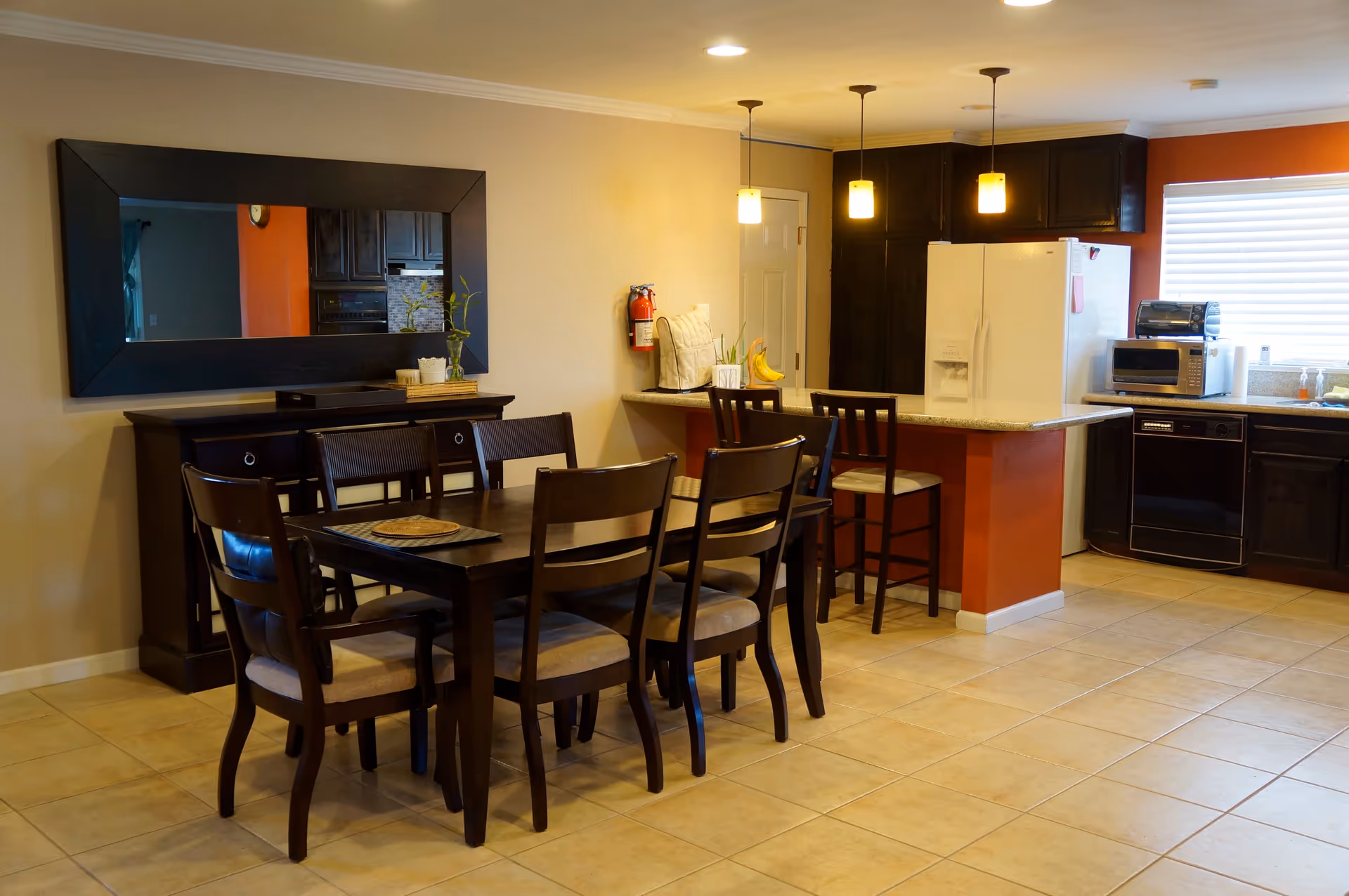 A dining area and kitchen in a senior living facility. The dining area has a dark wooden table with six chairs. On the wall above a sideboard is a large rectangular mirror. The kitchen features a white refrigerator, dark wooden cabinets, a countertop with two bar stools, a microwave, a toaster oven, and a dishwasher. The room has tiled flooring and warm lighting from three pendant lights above the counter.