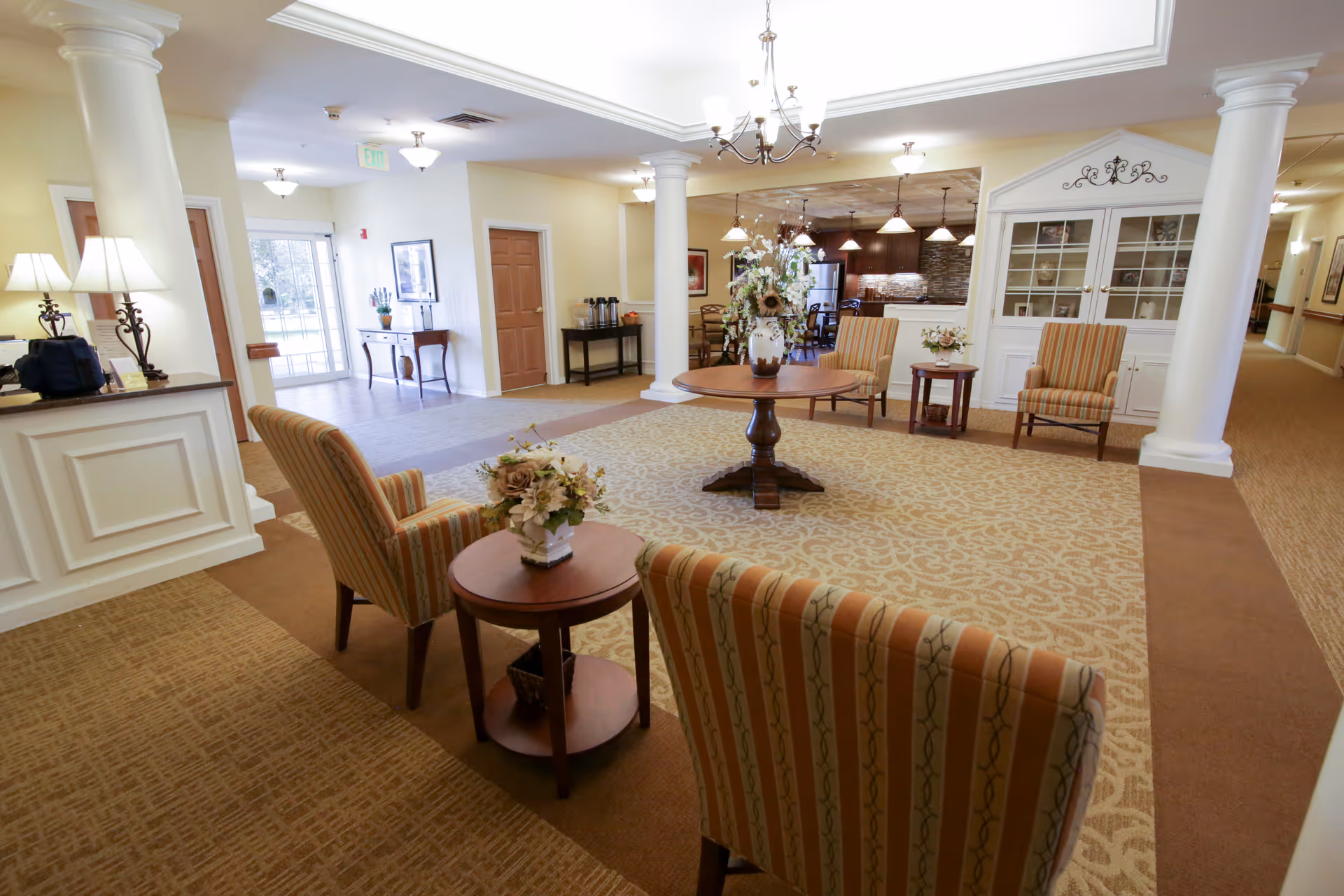 A bright and spacious common area in a senior living facility featuring a seating arrangement with striped upholstered chairs and small wooden tables with floral centerpieces. The room has large white columns, a chandelier, and a carpeted floor with a patterned rug. In the background, there is a kitchen area with pendant lights and wooden cabinetry, as well as a hallway and an entrance door with glass panels.