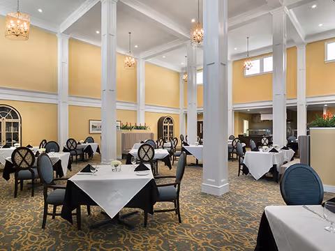 A spacious dining room with high ceilings, yellow walls, and white columns. The room is furnished with multiple tables covered with white tablecloths and black underlays, each set with black napkins and glassware. Elegant chandeliers hang from the ceiling, and the carpet has a decorative pattern. There are large windows and arched doorways in the background.