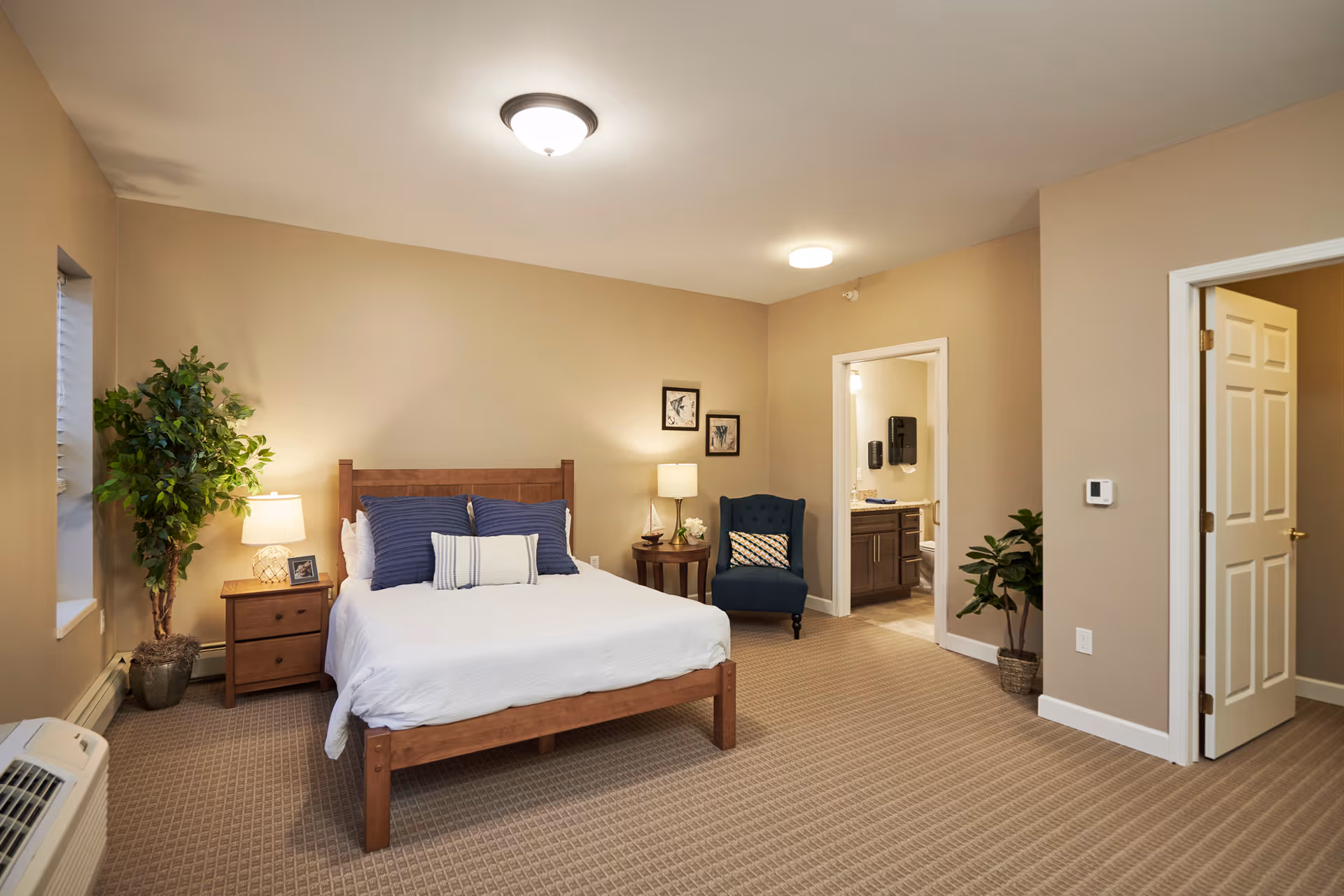 A well-lit bedroom in a senior living facility featuring a wooden bed with white bedding and blue pillows, a wooden nightstand with a lamp and picture frame, a blue upholstered chair with a decorative pillow, two framed pictures on the wall, a potted plant near the window, and an open door leading to a bathroom.