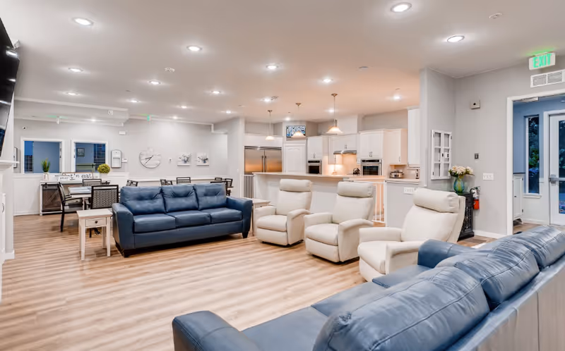 Spacious open living area with blue sofas and cream recliners facing a modern kitchen and dining area.