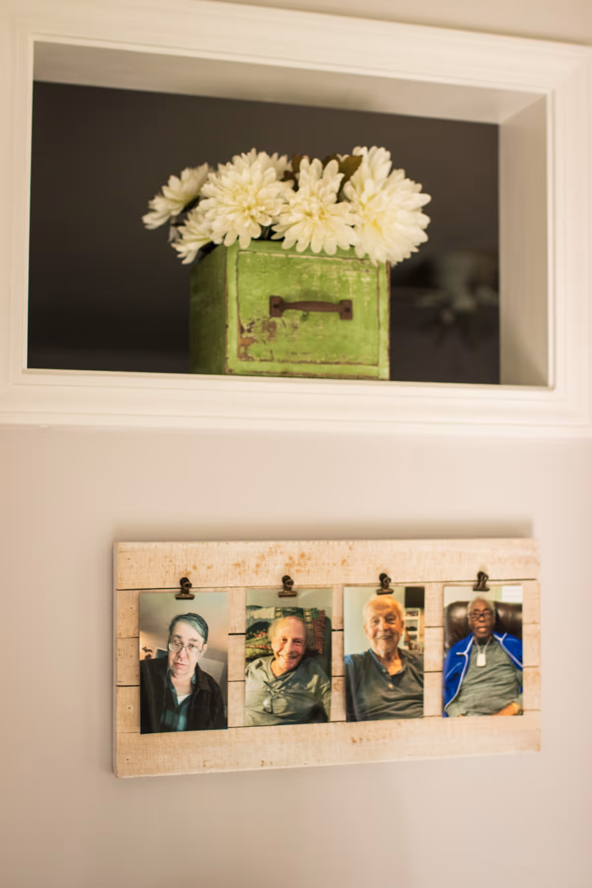 Interior wall display with a recessed niche holding a green box of white flowers above a wooden photo board showing four portrait photos.