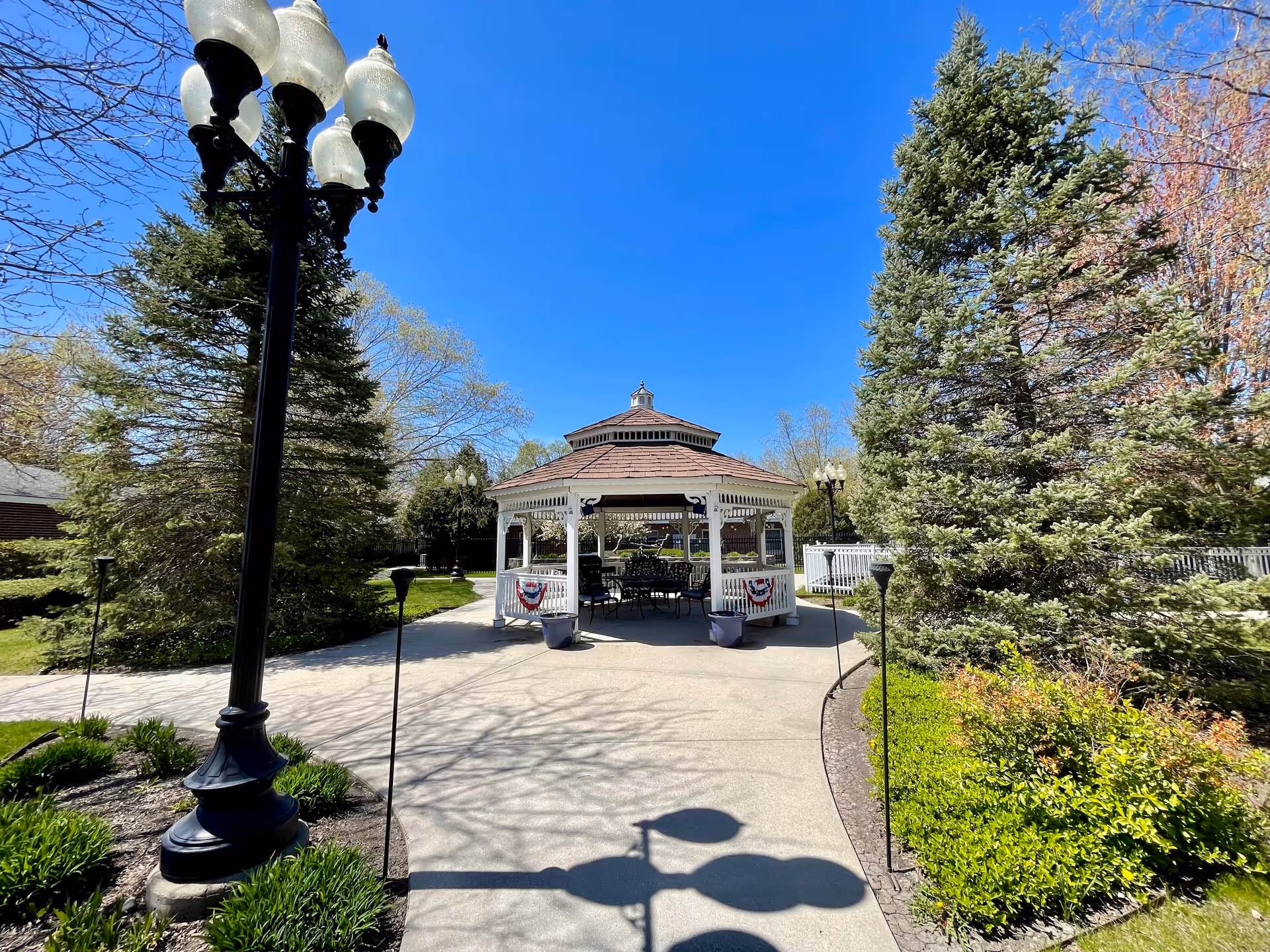 A white gazebo with a brown roof is situated at the end of a paved pathway surrounded by green bushes and tall trees under a clear blue sky. There are black metal chairs and tables inside the gazebo. A black street lamp with four glass globes stands on the left side of the pathway.