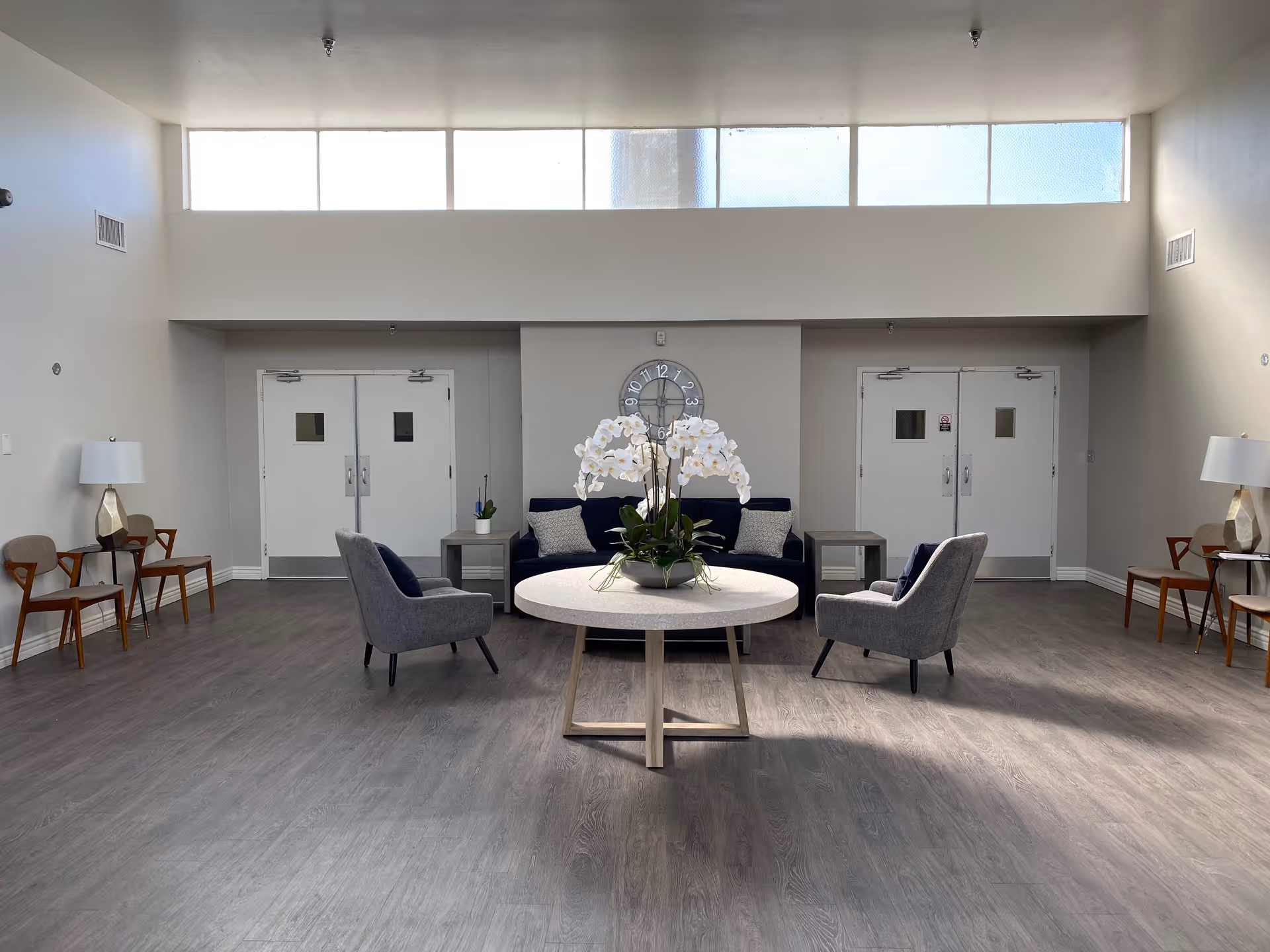 A bright, spacious lounge with a round table topped with white orchids, armchairs, a sofa, and double doors in the background.