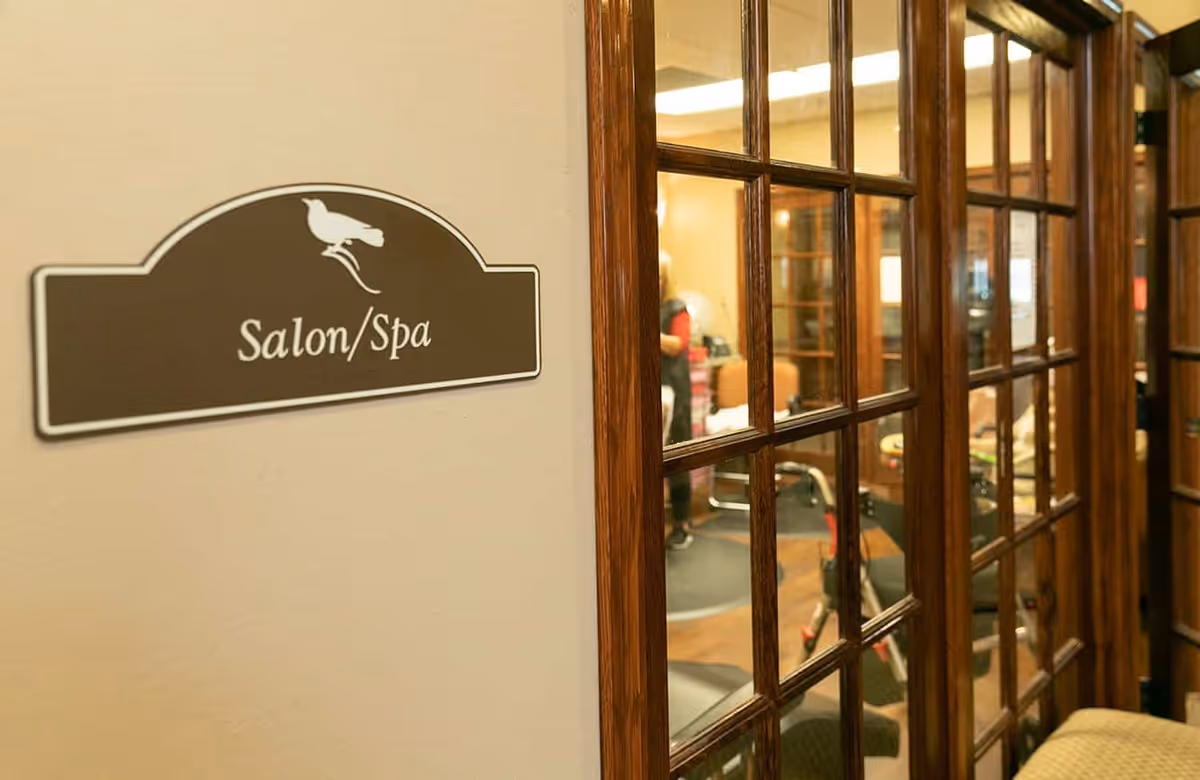 A close-up view of a wall sign that reads 'Salon/Spa' with a bird graphic above the text. To the right, there are wooden-framed glass doors revealing a glimpse of the interior salon/spa area with chairs and a person inside.